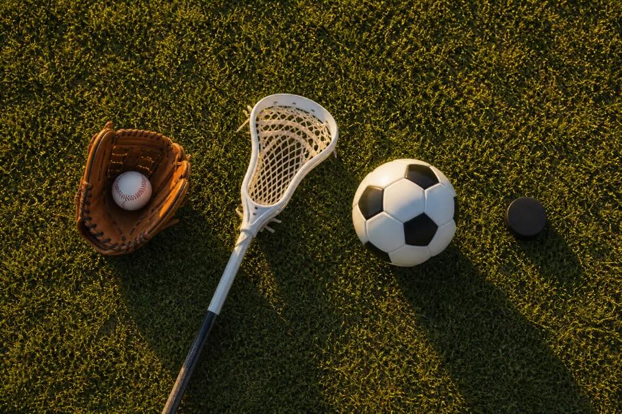 Overhead flat-lay of youth sports equipment on a grassy field, featuring a baseball glove with a ball, a lacrosse stick, a soccer ball, and a hockey puck arranged in a horizontal layout. Warm golden-hour sunlight casts long shadows across the scene, with no people or text present.